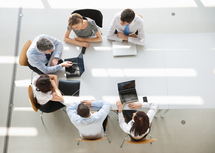 Aerial view of six people sitting around a white table in a modern office. They are engaged in a discussion with laptops open. Sunlight streams through windows, casting lines on the floor.