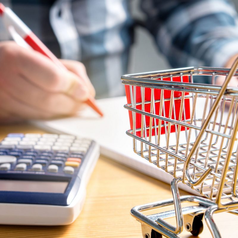 A person is writing on a notepad next to a calculator on a wooden table. In the foreground, there is a small shopping cart. The scene suggests calculating expenses or budgeting.