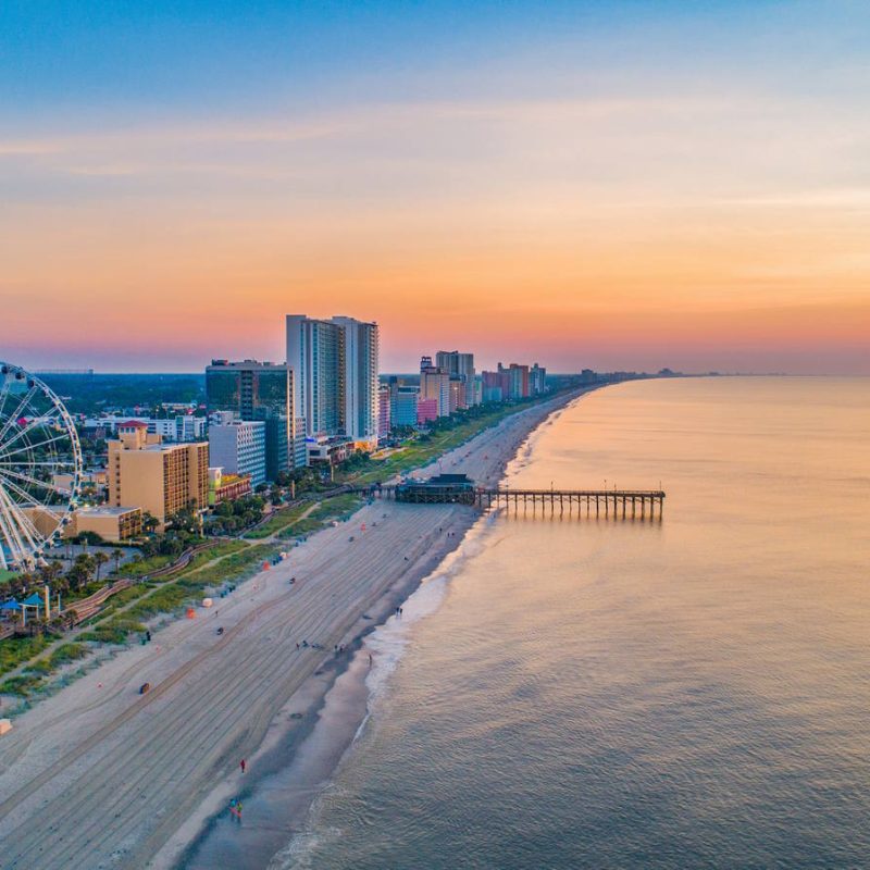 Aerial view of a coastal city at sunset with a sandy beach and calm ocean. A Ferris wheel and tall buildings line the shore, with a pier extending into the water. The sky is colorful with hues of orange and pink as the sun sets on the horizon.