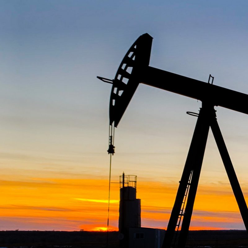 Silhouette of an oil pump jack against a vibrant sunset sky. The horizon glows with shades of orange and yellow, contrasting with the dark outline of the machinery.