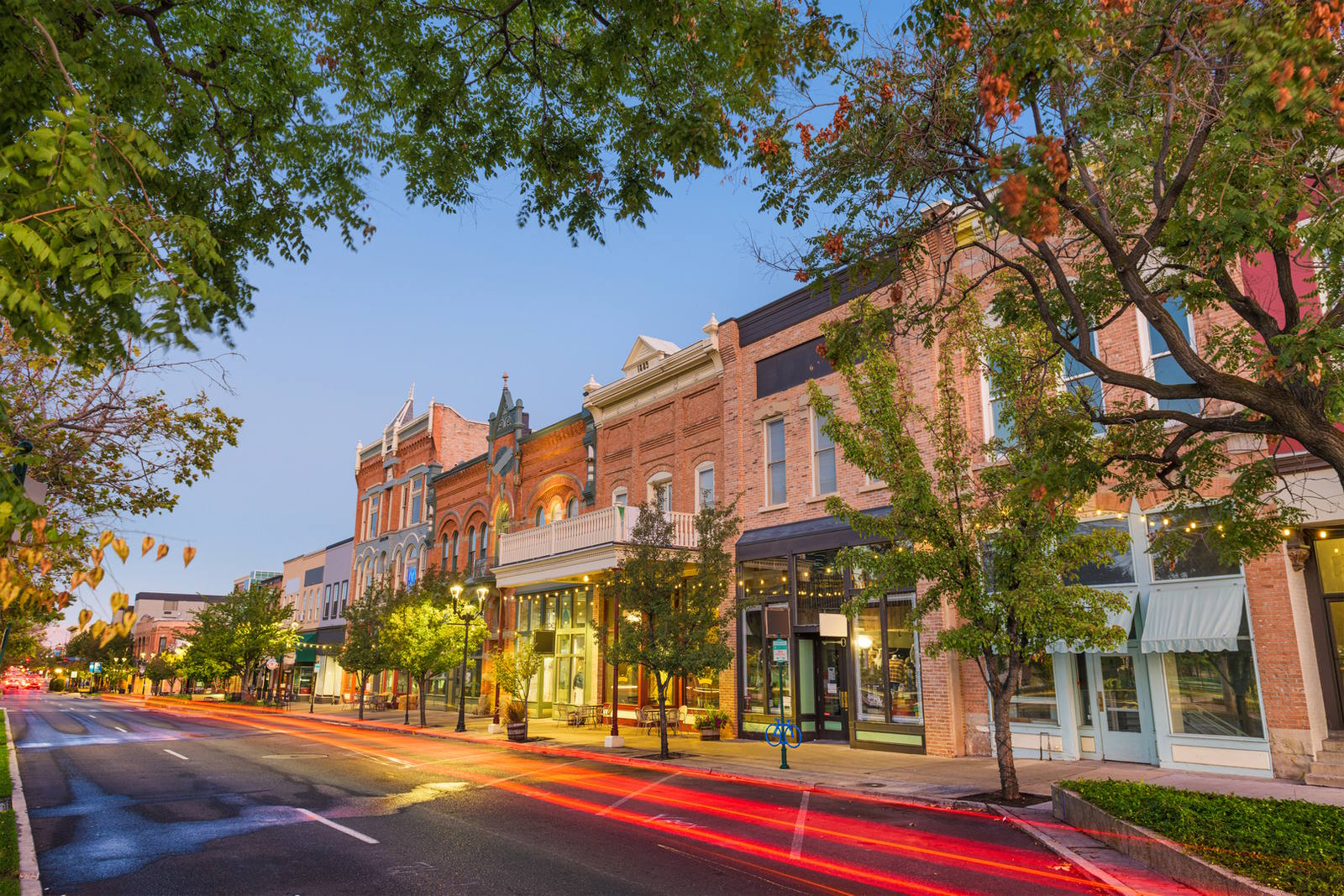 A scenic view of a small town main street at dusk. Historic brick buildings line the street, with trees and glowing streetlights. The sky is clear, and light trails from passing cars create a dynamic effect on the road.