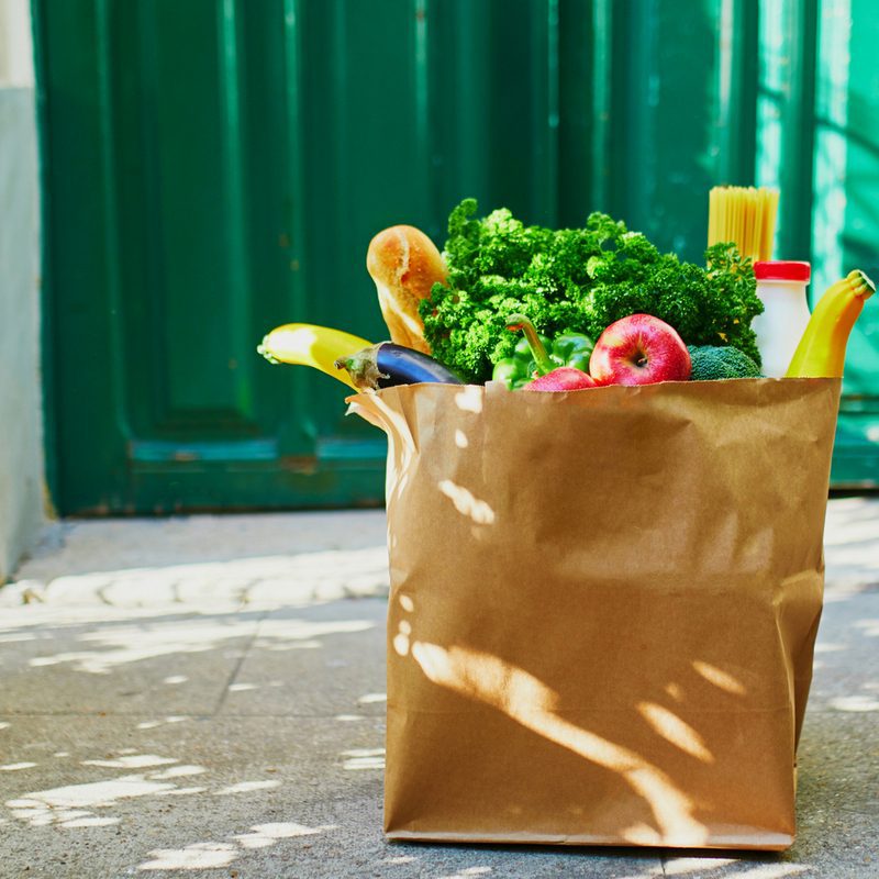 A brown paper bag filled with fresh groceries, including bananas, parsley, an apple, baguette, and pasta, sits on a sunlit concrete doorstep with a green door in the background. Shadow patterns from nearby foliage are visible on the ground.