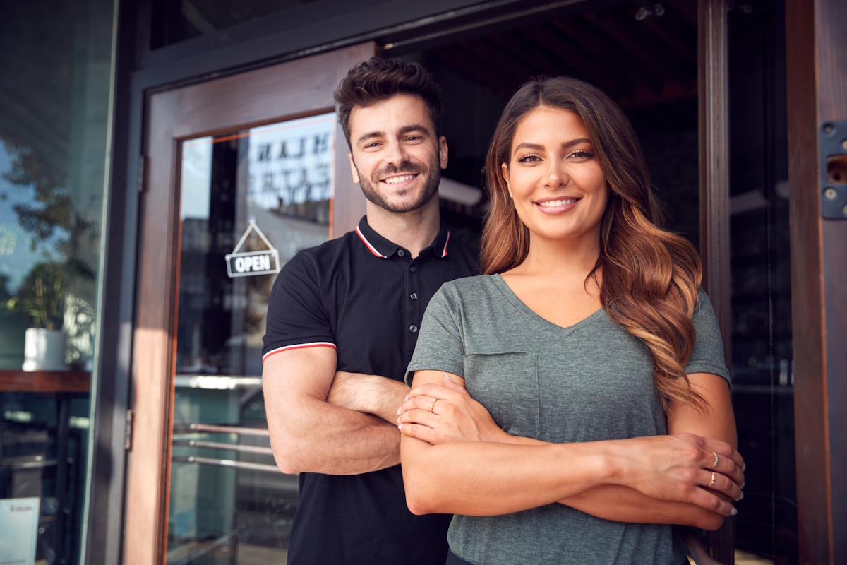 A man and woman stand smiling in front of a glass door with an Open sign. The man is in a black polo shirt, and the woman is in a gray shirt. They appear to be outside a shop or café, standing close to each other.