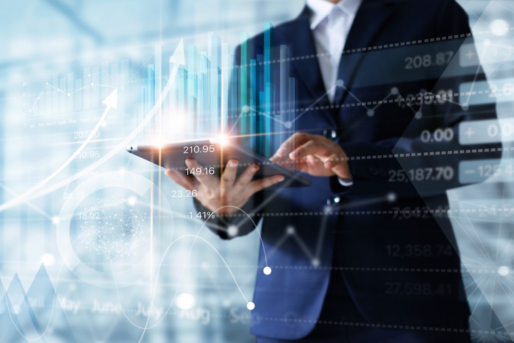 A person in a suit uses a tablet against a backdrop of digital graphs and charts. Bright lines and numbers display stock or financial data, giving a sense of analytics and technology in finance.