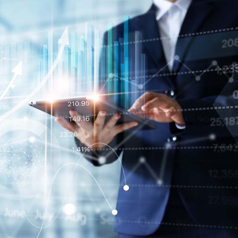 A person in a suit uses a tablet against a backdrop of digital graphs and charts. Bright lines and numbers display stock or financial data, giving a sense of analytics and technology in finance.