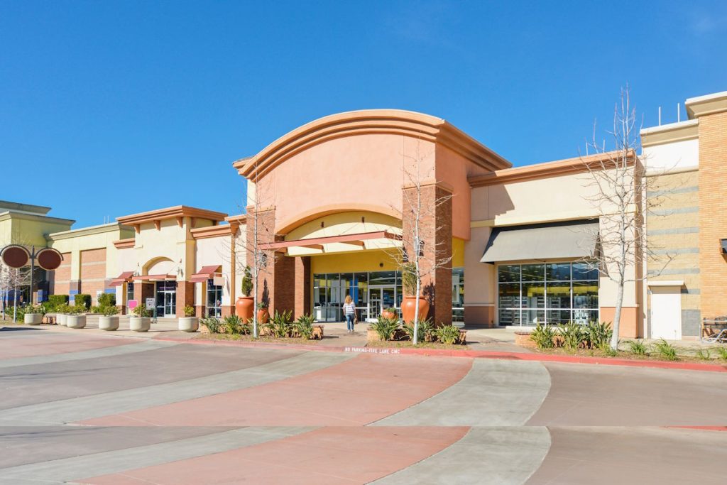 A shopping center with multiple storefronts featuring large glass windows and awnings. The buildings are a mix of warm colors like orange and beige. The wide paved walkway is in front, with a clear blue sky above.
