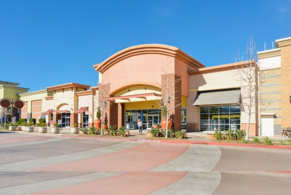 A shopping center with multiple storefronts featuring large glass windows and awnings. The buildings are a mix of warm colors like orange and beige. The wide paved walkway is in front, with a clear blue sky above.