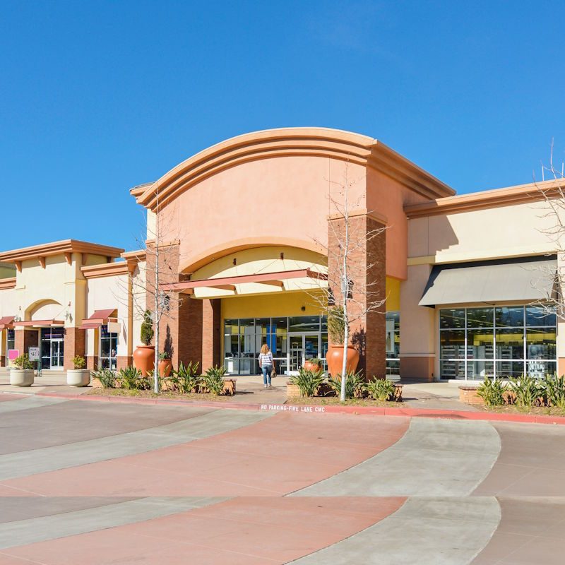 A shopping center with multiple storefronts featuring large glass windows and awnings. The buildings are a mix of warm colors like orange and beige. The wide paved walkway is in front, with a clear blue sky above.