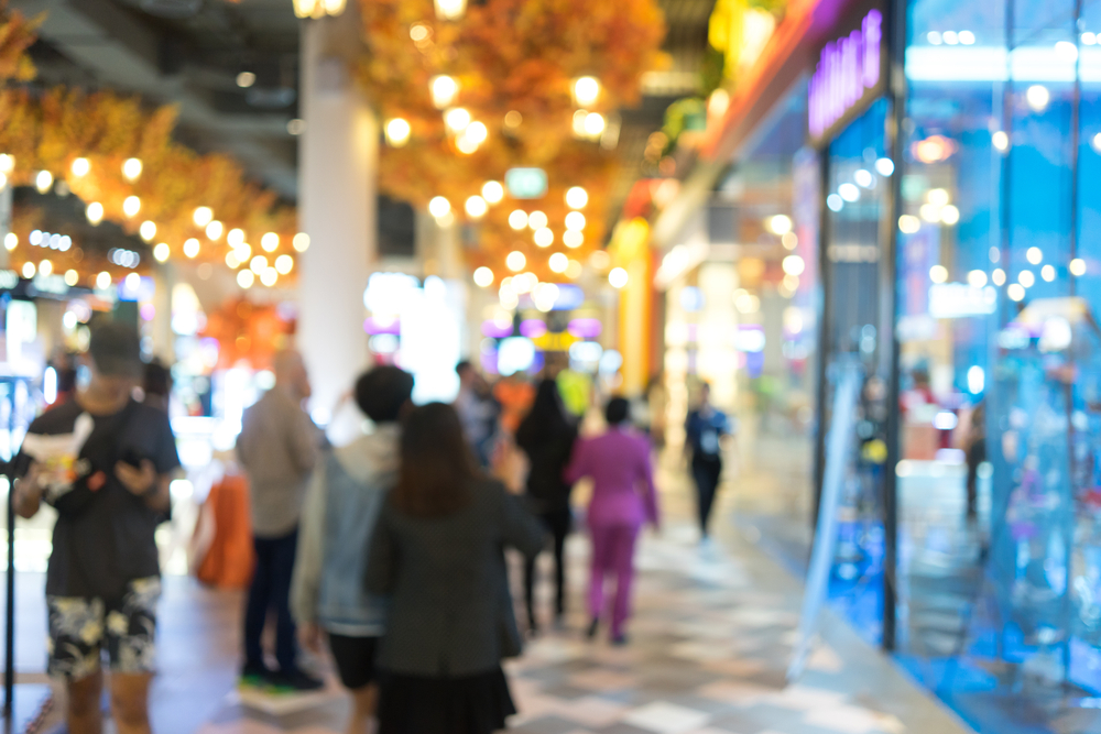 A blurred image of people walking in a brightly lit shopping mall, with colorful stores and glowing lights creating a lively atmosphere. The focus is on the bustling crowd rather than specific details.