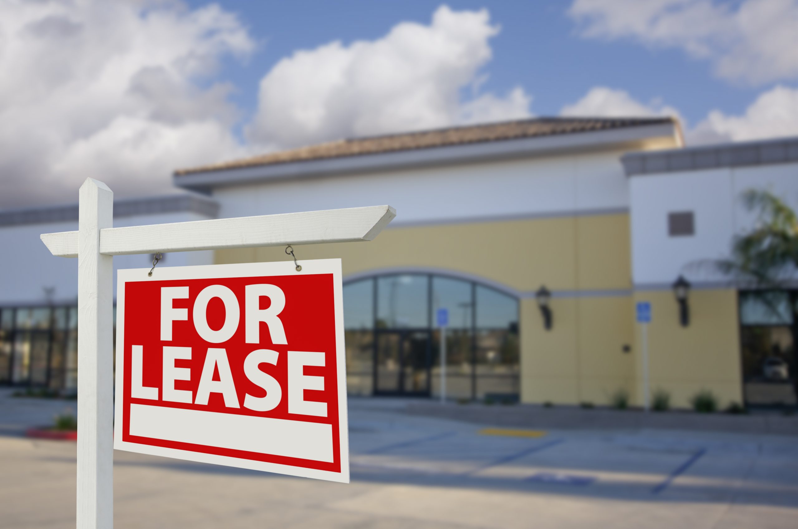 A For Lease sign is prominently displayed in front of a vacant commercial building. The building has a cream and yellow facade, large glass windows, and a tiled roof, set against a partly cloudy sky.