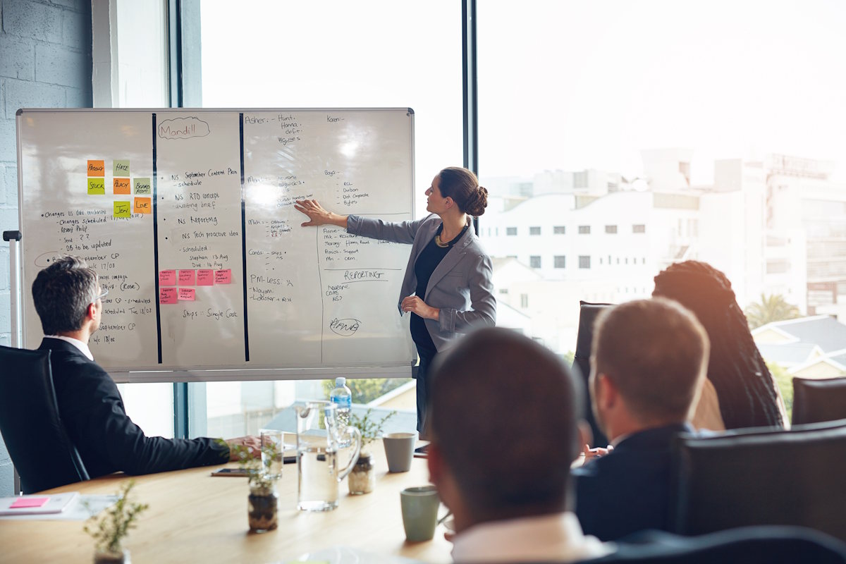 A person stands in a conference room, pointing at a whiteboard filled with notes and diagrams. Three colleagues sit around a table, listening attentively. Large windows in the background reveal an urban cityscape.
