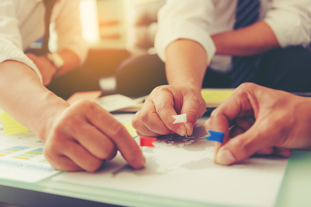 Three people in business attire are gathered around a map on a table. They are using small colored flags to mark points on the map, suggesting planning or strategy. The scene is well-lit and suggests collaboration.