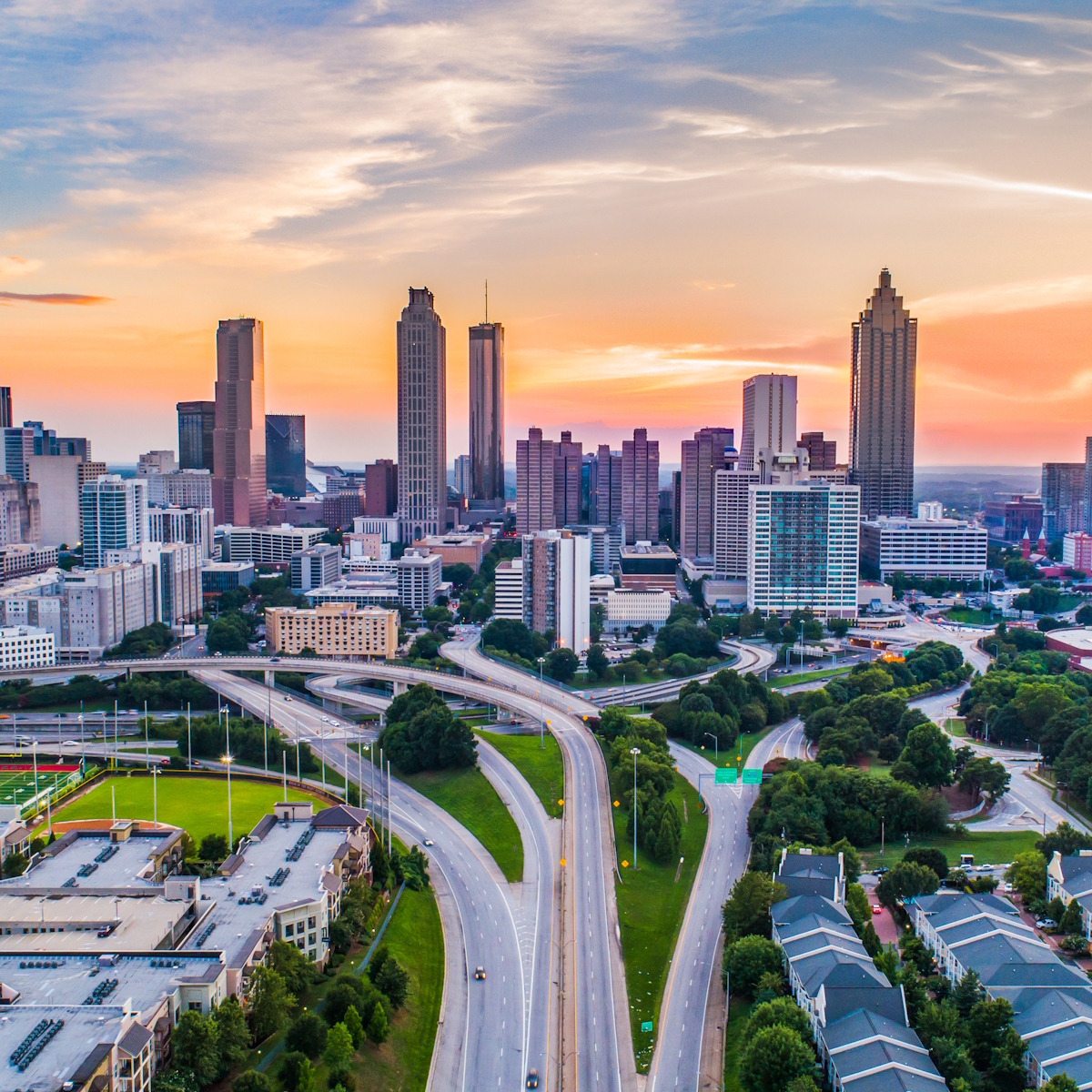 Aerial view of Atlanta, GA at sunset, featuring tall skyscrapers and a network of highways converging towards the city center. The sky displays vibrant hues of orange, pink, and blue, with scattered clouds adding texture to the scene.