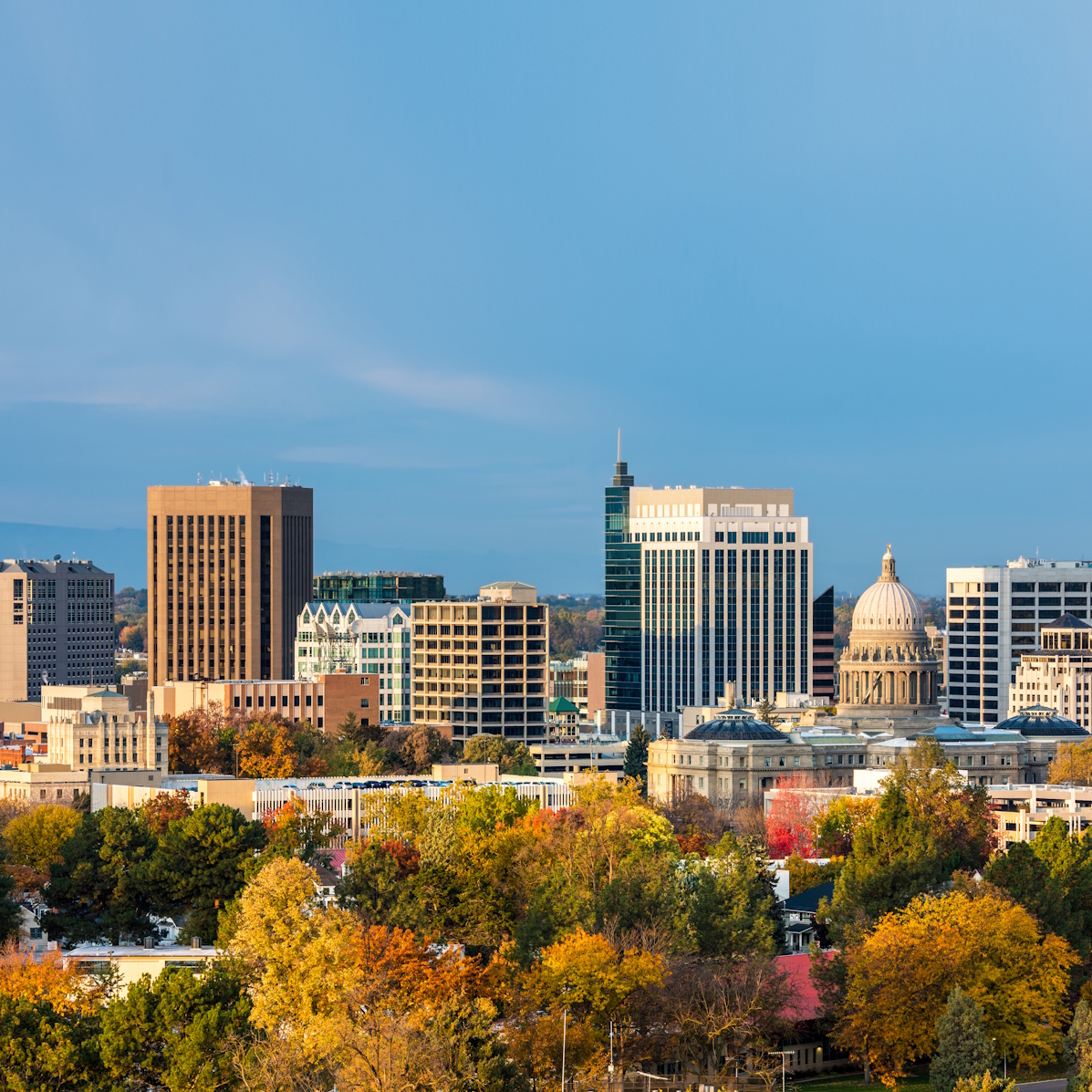 Skyline of Boise, Idaho, including the domed capitol building. The foreground features trees displaying autumn colors, and the sky is clear with soft, golden sunlight.