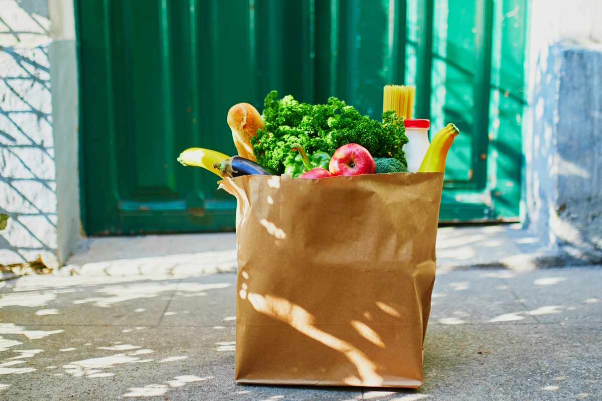 In the highly competitive grocery industry, companies rely on proven site selection and forecasting methods. A brown paper bag filled with groceries, including bananas, an apple, a baguette, broccoli, kale, a bottle, and a pack of spaghetti.