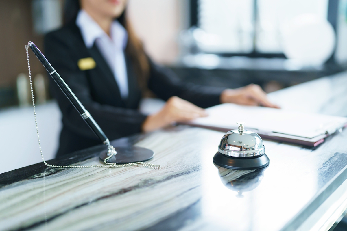 A hotel reception desk with a service bell and a pen on a chain in the foreground. A person in a uniform jacket is blurred in the background, holding a clipboard. The counter has a marbled surface.