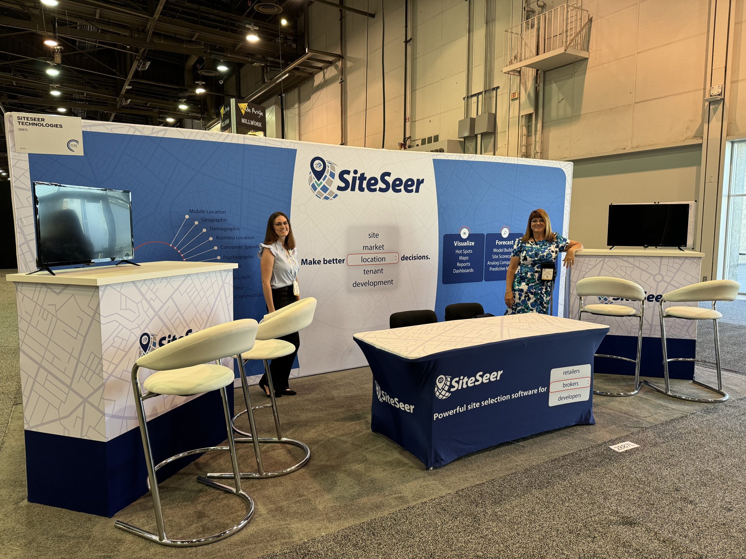 Two people stand at a trade show booth displaying SiteSeer branding. The booth features a blue and white color scheme, maps on the backdrop, and tables with promotional materials. Monitors and a seating area are also visible.