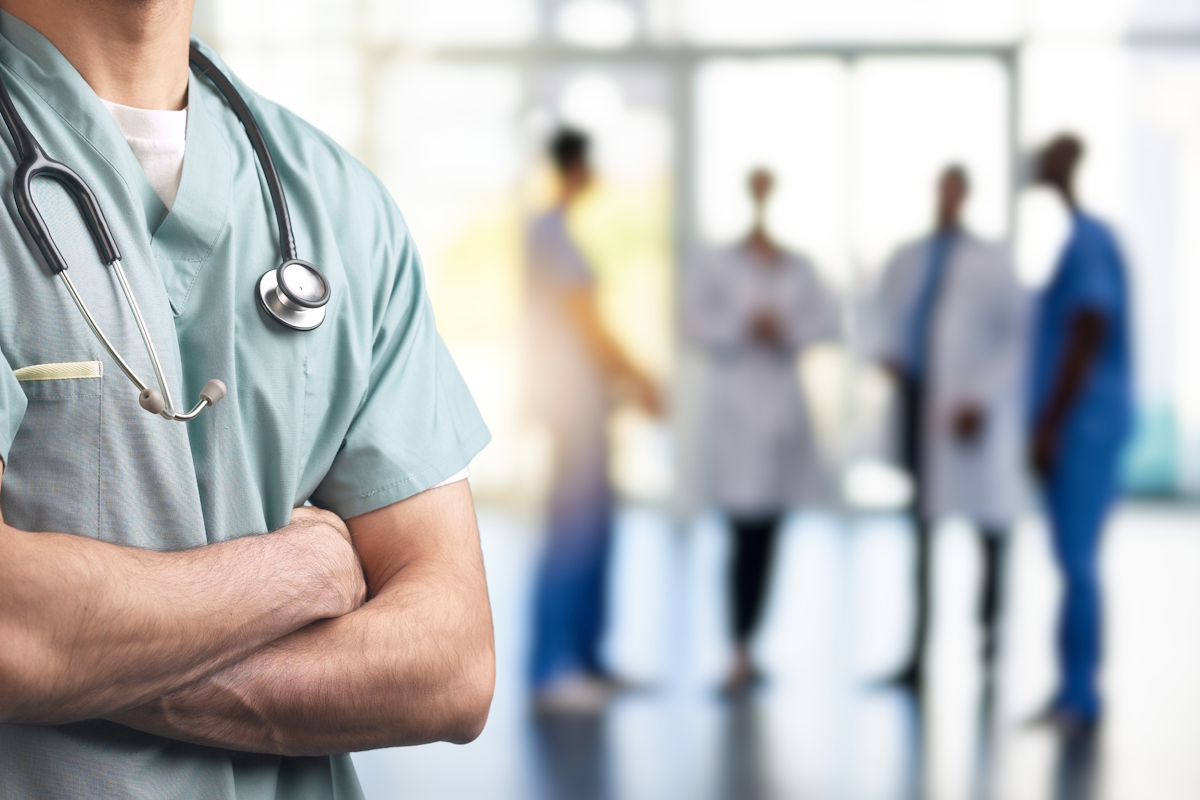 A medical professional in green scrubs, with arms crossed and a stethoscope around the neck, stands in focus. In the blurred background, several healthcare workers in white coats and blue scrubs are conversing in a bright, modern hospital setting.